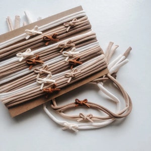May include: A set of 18 nude and brown stretchy headbands with suede bows in brown, beige, and white. The headbands are arranged on a brown card.