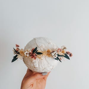 Puede incluir: Una corona floral crema con flores secas en tonos rosa, blanco y verde, acentuada por una cinta amarilla suave. La corona est&aacute; dise&ntilde;ada para un beb&eacute; o ni&ntilde;o, perfecta para una sesi&oacute;n de fotos o una ocasi&oacute;n especial.