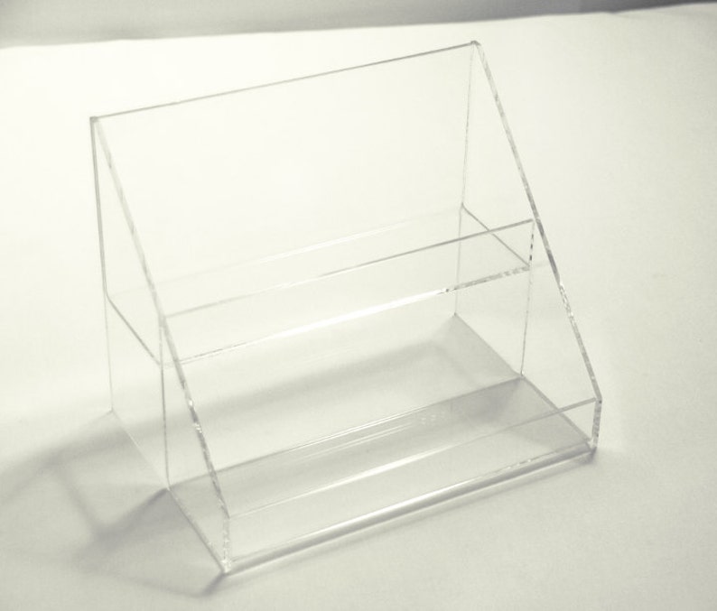 K&ouml;nnte beinhalten: Ein zweistufiger Acryl-Pr&auml;sentationsst&auml;nder mit schr&auml;ger Vorderseite. Der St&auml;nder ist aus klarem Acryl gefertigt und hat ein modernes Design.