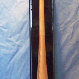 May include: A baseball bat and ball displayed in a clear case. The baseball sits atop the bat's handle. The bat is light brown, with a rounded handle and a wider barrel. The baseball has red stitching, and the case is rectangular.