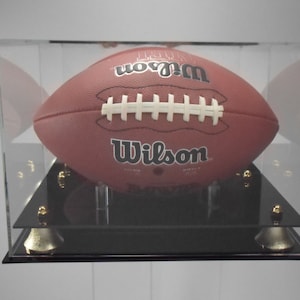 Puede incluir: Un balón de fútbol de cuero marrón con cordones blancos y la palabra "Wilson" impresa en él se muestra en una vitrina de acrílico transparente con una base negra. La vitrina tiene patas de metal doradas.