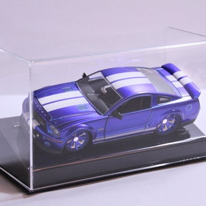 May include: A blue and white model car in a clear acrylic display case. The car has white racing stripes and a black base.