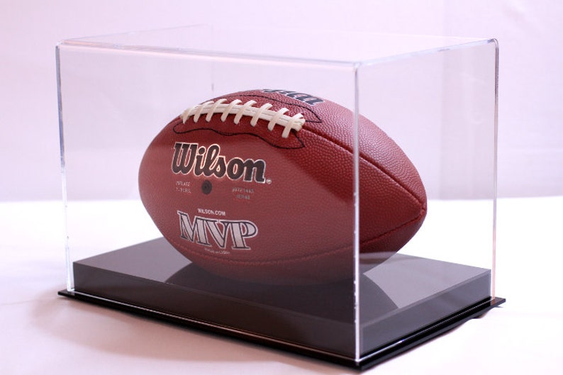May include: A brown Wilson MVP football inside a clear acrylic display case. The football has white laces and the Wilson logo. The display case sits on a black base, providing a clean presentation for sports memorabilia.