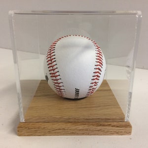 May include: A white baseball with red stitching is displayed in a clear acrylic case with a wooden base.
