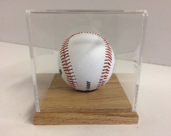 Könnte beinhalten: Ein weißer Baseball mit roten Nähten wird in einem durchsichtigen Acryl-Gehäuse mit einem Holzsockel präsentiert.