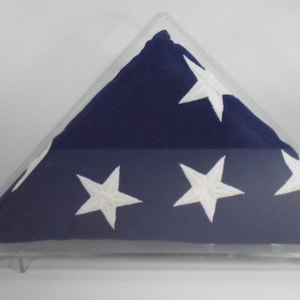 Könnte beinhalten: Eine gefaltete blaue Flagge mit weißen Sternen, präsentiert in einem transparenten Acrylgehäuse. Die Flagge ist dreieckig gefaltet, wobei die Sterne auf der Oberseite sichtbar sind. Das Gehäuse wird von kleinen, transparenten Ständern getragen.