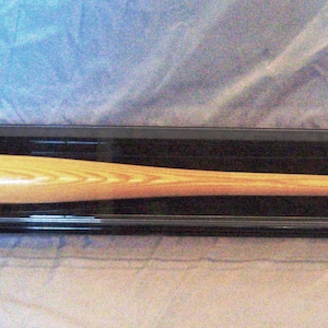 May include: A wooden baseball bat displayed in a clear acrylic case with a black base. The bat is light brown with a natural wood grain.