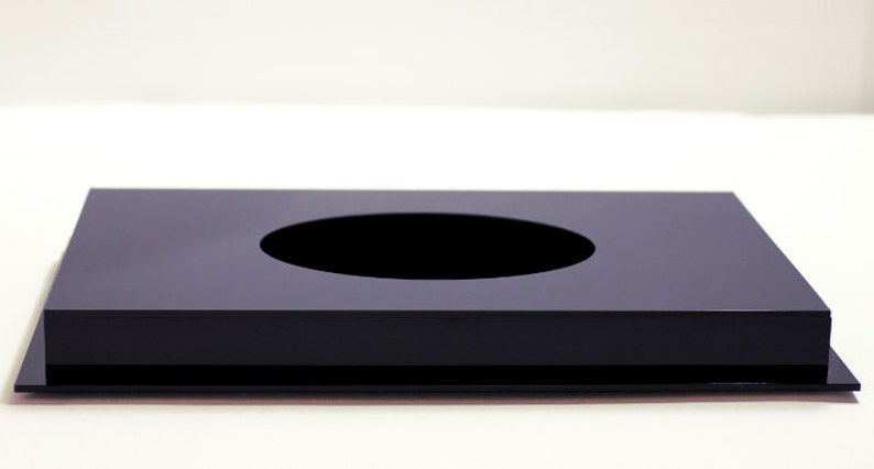 May include: A black, rectangular, modern tissue box cover with a circular opening in the center. The tissue box cover is made of a glossy material and sits on a flat base.