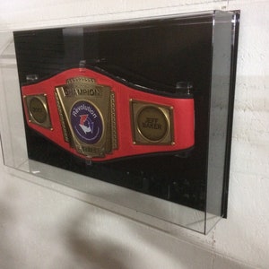K&ouml;nnte beinhalten: Ein roter Meisterschaftsg&uuml;rtel mit goldenen Akzenten und den Worten "Champion" und "Revolution Title" darauf. Der G&uuml;rtel hat auch eine goldene Plakette mit dem Namen "Jeff Baker" darauf.