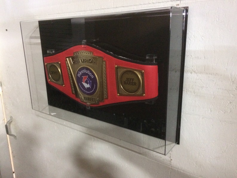 K&ouml;nnte beinhalten: Ein roter Meisterschaftsg&uuml;rtel mit goldenen Akzenten und den Worten "Champion" und "Revolution Title" darauf. Der G&uuml;rtel hat auch eine goldene Plakette mit dem Namen "Jeff Baker" darauf.