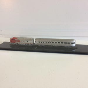 May include: A model train set featuring a red and silver locomotive and a silver passenger car, displayed in a clear acrylic case. The train cars sit on a black base with simulated tracks, ideal for collectors or hobbyists.