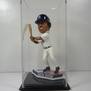 May include: A clear acrylic display case with a black base, showcasing a bobblehead figurine of a baseball player wearing a white and blue uniform with a blue baseball cap with a white "D" on it. The base of the bobblehead features the text "Austin Jackson".