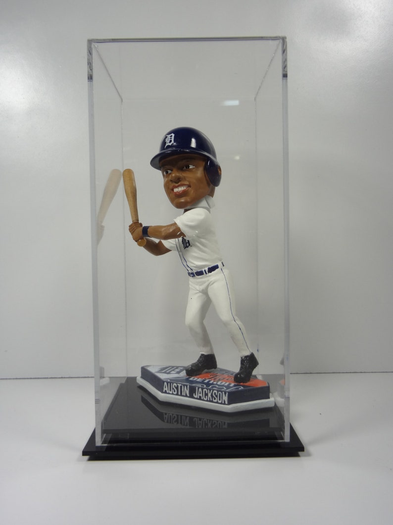 May include: A clear acrylic display case with a black base, showcasing a bobblehead figurine of a baseball player wearing a white and blue uniform with a blue baseball cap with a white "D" on it. The base of the bobblehead features the text "Austin Jackson".
