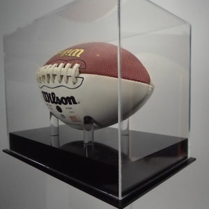 May include: A clear acrylic display case with a black base, showcasing a brown and white football with the word "Wilson" printed on it.