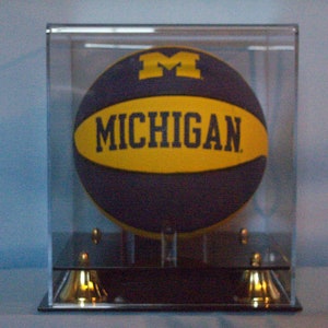 Pode incluir: Uma bola de basquete azul marinho e amarela com a palavra "MICHIGAN" e um logotipo "M" amarelo, exibida em uma vitrine de acrílico transparente. A vitrine tem uma base preta com detalhes dourados.