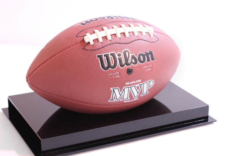 May include: A brown leather Wilson MVP football with white laces, displayed on a black rectangular stand. The football has the Wilson logo and the text "MVP" printed on it. The football is inflated to 7-9 lbs.