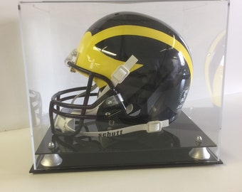 May include: A black and yellow football helmet with a white face mask and a white strap with the word "Schutt" on it. The helmet is displayed in a clear acrylic case with a black base.