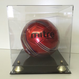 Puede incluir: Un balón de fútbol rojo y blanco con la palabra "mitre" impresa en él se muestra en una vitrina de acrílico transparente con una base negra.