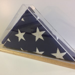 Könnte beinhalten: Eine gefaltete amerikanische Flagge in einem transparenten Acrylgehäuse mit Holzsockel. Die Flagge ist blau mit weißen Sternen und wird in einem dreieckigen Gehäuse präsentiert.