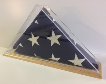 May include: A folded American flag displayed in a clear acrylic case with a wooden base. The flag is blue with white stars and is encased in a triangular display case.