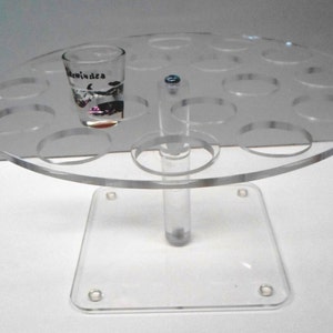 May include: A clear acrylic shot glass holder with a round top and a square base. The holder has 12 slots for shot glasses. A shot glass is in one of the slots.