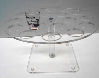 May include: A clear acrylic shot glass holder with a round top and a square base. The holder has 12 slots for shot glasses. A shot glass is in one of the slots.