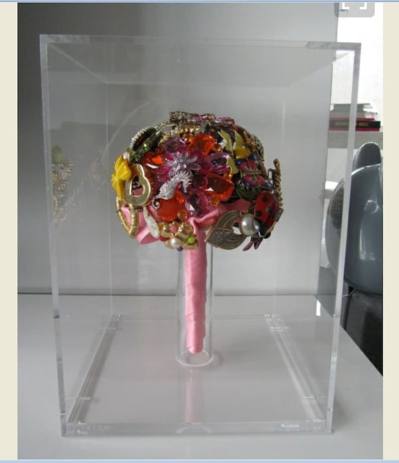 May include: A clear acrylic box with a pink ribbon bouquet made of various colorful brooches, rhinestones, and other jewelry.