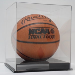 Pode incluir: Uma bola de basquete de couro marrom com marcas pretas, incluindo o texto "Rawlings" e "NCAA Final Four", exibida em uma vitrine de acrílico transparente com uma base preta.