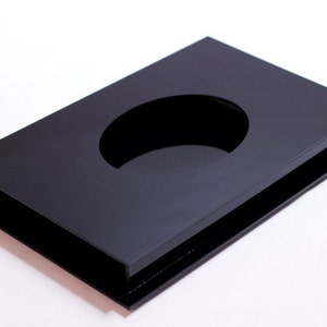 May include: A rectangular, black acrylic tray with a large oval cutout in the center. The tray has a raised border and sits on a slightly larger base, creating a layered effect. The glossy surface reflects light.