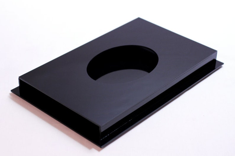 May include: A rectangular, black acrylic tray with a large oval cutout in the center. The tray has a raised border and sits on a slightly larger base, creating a layered effect. The glossy surface reflects light.