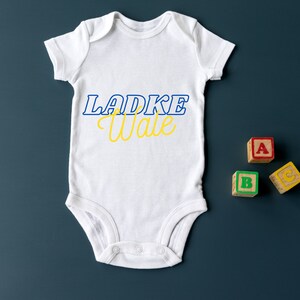 May include: White baby onesie with blue and yellow lettering that says "Ladke Wale". Three wooden blocks with the letters A, B, and C are on a blue background.