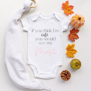 May include: White baby onesie with the text "If you think I'm cute you should see my Masi" in gray and pink lettering. The onesie is surrounded by fall-themed decorations, including small pumpkins and fall leaves.