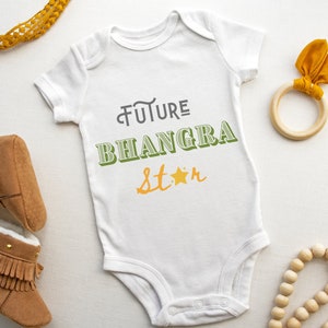 May include: White baby onesie with the text "FUTURE BHANGRA star" in green and yellow lettering. The onesie is surrounded by brown baby booties, a wooden ring with a yellow bow, and a wooden bead necklace.