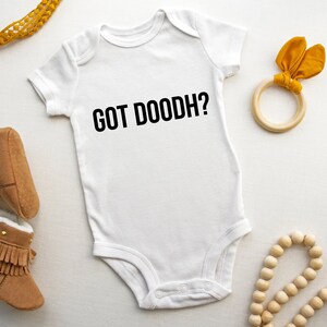 May include: White baby onesie with the text "GOT DOODH?" in black. The onesie is surrounded by brown baby booties, a wooden ring with yellow fabric, and a wooden bead necklace with a brown tassel.