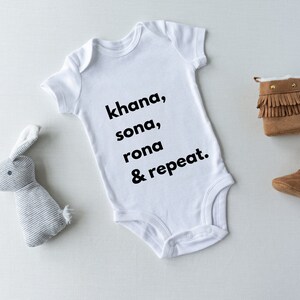 May include: White baby onesie with the text "khana, sona, rona & repeat."  A gray stuffed bunny and two brown suede baby booties with fringe are on a white background.