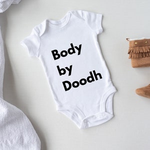 May include: White baby onesie with black text that reads "Body by Doodh".  Brown baby booties with fringe and gold accents are on a white surface.