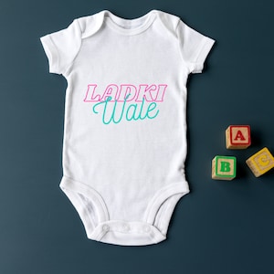 May include: White baby onesie with the text "Ladki Wale" in pink and teal lettering. Three wooden blocks with the letters A, B, and C are on a dark blue background.