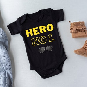 May include: Black baby onesie with yellow text that reads "HERO NO 1" and a pair of sunglasses printed on the front.