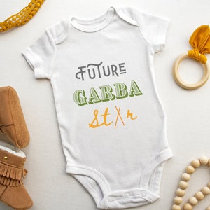 May include: White baby onesie with the text "FUTURE GARBA STAR" in green and orange lettering. The onesie is surrounded by brown baby booties, a wooden ring with a yellow fabric bow, and a wooden bead necklace.