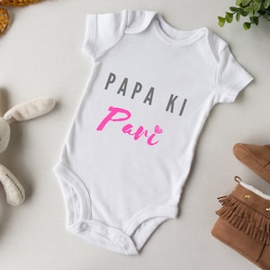 May include: White baby onesie with the text "Papa Ki Pari" in grey and pink lettering. The onesie is surrounded by a white bunny, brown booties, and a gold ribbon.