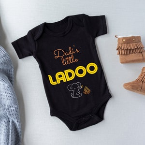 May include: Black baby onesie with the text "Dadi's little LADOO" in yellow and orange letters. A small elephant illustration is below the text. The onesie is next to a pair of brown baby booties with fringe.