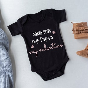 May include: Black baby onesie with the text "Sorry boys my Papa's my valentine" in white with pink hearts. The onesie is laying on a white surface with a blue blanket and brown baby booties.
