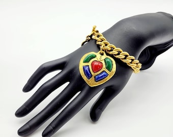 Puede incluir: Un brazalete de cadena dorada con un gran dije en forma de corazón. El dije está hecho de metal dorado y tiene acentos de esmalte rojo, azul y verde.
