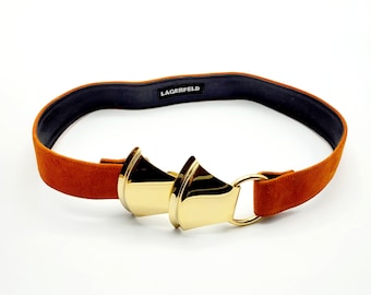 May include: A brown suede belt with a gold metal buckle. The buckle is a double-sided design with a triangular shape. The belt is labeled "LAGERFELD".