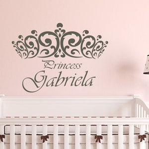 Może przedstawiać: R&oacute;żowa ściana z ozdobną koroną w kolorze szarym i napisem "Princess Gabriela" w ozdobnym, pochylonym piśmie. Na pierwszym planie znajduje się białe ł&oacute;żeczko z białym materacem i brązowymi kokardkami.