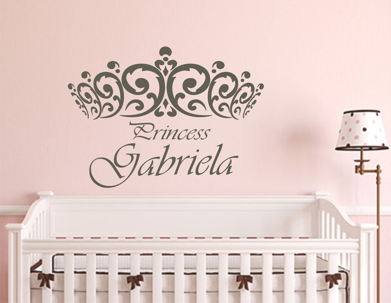 Może przedstawiać: R&oacute;żowa ściana z ozdobną koroną w kolorze szarym i napisem "Princess Gabriela" w ozdobnym, pochylonym piśmie. Na pierwszym planie znajduje się białe ł&oacute;żeczko z białym materacem i brązowymi kokardkami.