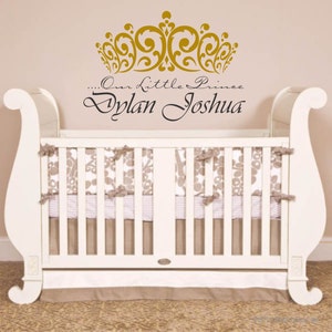 May include: A white crib with a decorative headboard and footboard. The crib is adorned with a floral patterned bedding and a skirt. Above the crib, a gold crown design and the text "...Our Little Prince Dylan Joshua" are displayed.