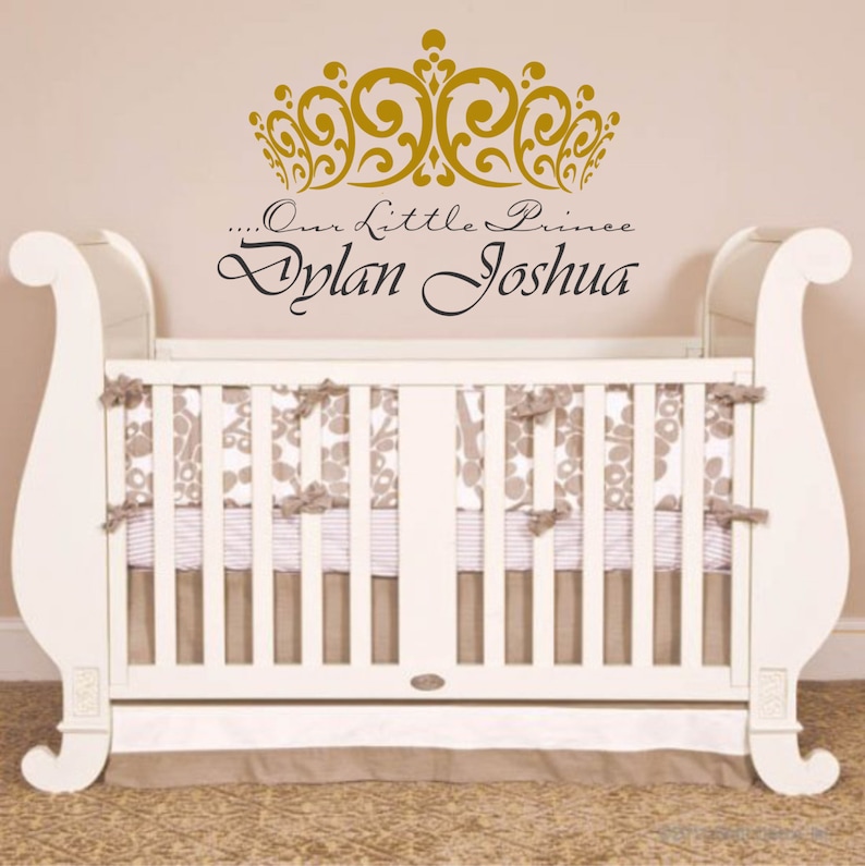 May include: A white crib with a decorative headboard and footboard. The crib is adorned with a floral patterned bedding and a skirt. Above the crib, a gold crown design and the text "...Our Little Prince Dylan Joshua" are displayed.