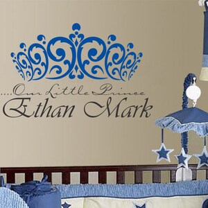 May include: A blue crown wall decal above the text "...Our Little Prince Ethan Mark" on a beige wall. A baby crib with blue bedding and a mobile with star-shaped ornaments is in the foreground.
