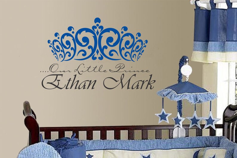 May include: A blue crown wall decal above the text "...Our Little Prince Ethan Mark" on a beige wall. A baby crib with blue bedding and a mobile with star-shaped ornaments is in the foreground.